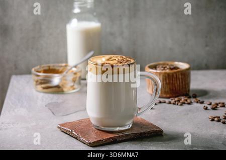 Dalgona schäumender Kaffee Trend koreanischer Drink Milchlatte mit Kaffeeschaum in Glasbecher, dekoriert mit gemahlenem Kaffee auf grauem Texturtisch. Zutaten oben. Kopierbereich Stockfoto