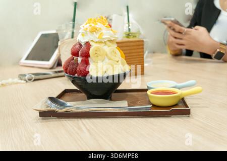 Ein Set koreanischer Bingsu in einer kleinen schwarzen Schüssel mit mehreren Früchten und roter Sauce mit süßer Kondensmilch in Mini-Plastikschüsseln in braunem Tablett Stockfoto