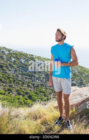 Erwachsener Mann, der auf einem Hügel an der Küste steht und eine blaue Flasche hält, ein blaues Hemd trägt, einen Kopierraum. Wanderer, Wanderweg, Küste, Meer, sonnig, robust, Gräser Stockfoto