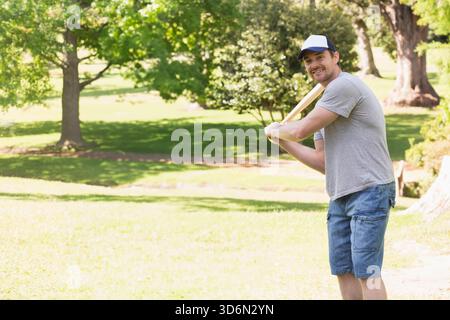 Mann in seinen 30ern, der hölzernen Baseballschläger hält, im Park steht, Trucker-Cap trägt, Kopierraum. Sportler, draußen, Sonnenlicht, Freizeit, Natur, Schatten Stockfoto
