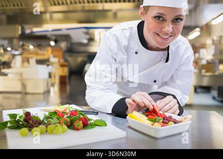 Weibliche Küchenchefin, die Obst auf weißem Geschirr anlegt und sich über die Theke in der Küche lehnt, mit weißem Jackenhut. Platte, Beschichtung, Edelstahl, Vorbereitung, Restaurant, cul Stockfoto