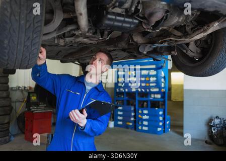 Männlicher Mechaniker, der das Fahrgestell auf hydraulischem Hubwerk überprüft, während er die Zwischenablage in der Werkstatt hält. Techniker, Overalls, blau, Garage, Werkstatt, Auspuff, Stockfoto