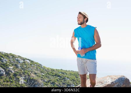 Mann, der auf einem felsigen Hügel steht, eine blaue Wasserflasche hält und ein blaues Hemd trägt, Kopierraum. Horizont, Meereslandschaft, Panorama, Wanderweg, Outdoor, erw Stockfoto