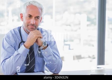 Reifer Arzt sitzt am Schreibtisch im modernen Büro und überprüft mit Stethoskop das Telefon anhand von Dokumenten. Arzt, Mann, Senior, sonnendurchflutete, luftige, Fenster, Citys Stockfoto