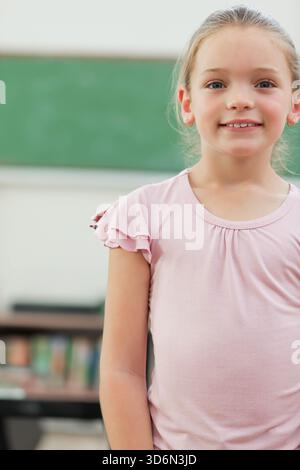 Schülerin, die im Klassenzimmer mit grüner Tafel steht und auf den Zuschauer blickt. Mädchen, Bildung, Lernen, Bücher, Schule, Pastell, neugierig Stockfoto