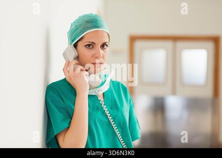 Chirurgin steht in grüner Schürze mit beigem schnurgebundenem Hörer im Krankenhaussaal, Maske hängt. Arzt, Medizin, Flur, Uniform, Kappe, Telefon Stockfoto