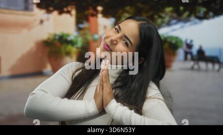 Die junge hispanische Frau lächelt mit Händen, die draußen in einer ruhigen Parklandschaft gefesselt sind, und trägt einen weißen Rollkragen mit sanftem Licht, das ihr Cheerfu hervorhebt Stockfoto