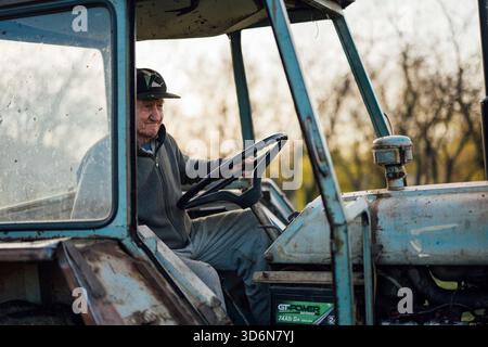 Älterer Landwirt, der im Herbst bei Sonnenuntergang auf einem Bauernhof einen Traktor fährt Stockfoto