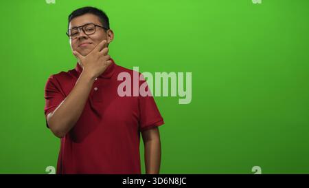 Junger Mann mit Hand auf dem Kinn, der in grünem Hintergrund im Studio posiert, mit rotem Polo und Brille; nachdenkliche Reflexion. Stockfoto