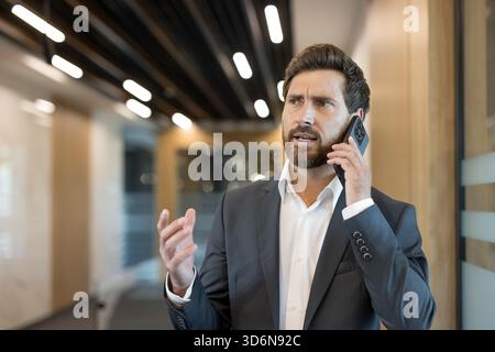 Geschäftsmann in Anzug und weißem Hemd, der ein ernsthaftes Gespräch auf einem Mobiltelefon führt, mit der Hand Gesten macht und besorgt aussieht, während er in einem modernen Flur steht Stockfoto