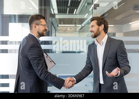 Zwei lächelnde Geschäftsleute in formeller Kleidung schütteln in einer modernen Büroumgebung die Hand und symbolisieren einen erfolgreichen Abschluss, eine Partnerschaft, ein Vertrauen und eine Zusammenarbeit Stockfoto