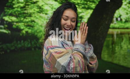 Frau mit Palmen zusammengedrückt nahe der Brust im grünen Park bei einem Baum, freundlich lächelnd und den Betrachter anschauend; Freude Dankbarkeit. Stockfoto