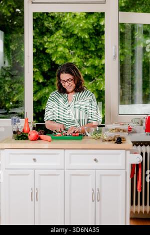 Midlife-Frau lächelt, während sie Gemüse an der Küchentheke hackt Stockfoto
