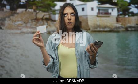Frau, die eine weiße Kreditkarte zwischen den Fingern hält und ein Smartphone in der anderen Hand am Strand mit einer verwunderten Stirn und leichtem Zucken; Zahlungsverwirrung. Stockfoto