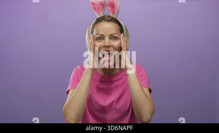 Frau hält die Wangen mit beiden Händen in einem lila Studio-Setting mit Hasenohren und rosa Hemd; Überraschung. Stockfoto