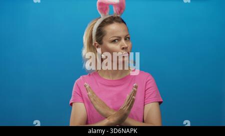 Die blonde Frau trägt ein Haarband mit Hasenohren und ein rosafarbenes Hemd kreuzt die Arme im hellen Studio; Ablehnung. Stockfoto