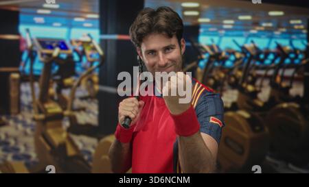 Mann, der Tennisschläger über die Schulter packt und geballte Faust in der Turnhalle mit Laufbändern und Heimtrainer in der Nähe pumpt; Siegesbestimmung. Stockfoto