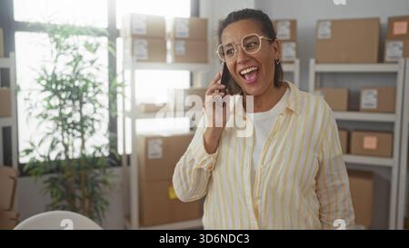 Die Frau hält das Smartphone am Ohr und lächelt zwischen Kartonschachteln auf den Regalen in einem Gebäude; der Besitzer eines kleinen Unternehmens ist fröhlich. Stockfoto