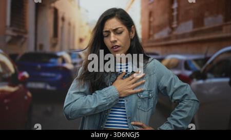 Frau, die den Mund mit der Hand bedeckt, auf einer belebten Straße neben geparkten Autos und alten Gebäuden hustet, Jeansjacke und gestreiftes Oberteil trägt; Atemnot di Stockfoto