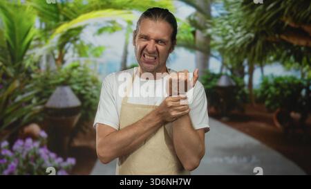 Mann trägt beige Schürzengrimaces, während er das Handgelenk auf einem von tropischem Grün gesäumten Parkweg hält; Unwohlsein. Stockfoto