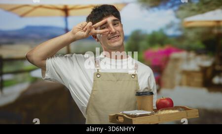Mann mit Schürze, der Holztablett mit Kaffeetasse, Gebäck und Apfel auf der Terrasse des Restaurants hält, die Hand zeigt ein Friedenszeichen vor Augen; freundlicher Service. Stockfoto