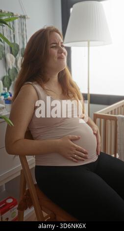 Junge kaukasische Frau, die auf Holzstuhl sitzt und schwangeren Bauch hält und im Kinderzimmer grimmt; Unwohlsein. Stockfoto