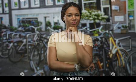 Frau mit Hand am Kinn auf dem Fahrradparkplatz auf der Straße, trägt Armbänder und Jeans und steht mit einem Arm über der Taille; durchdachtes Vertrauen. Stockfoto