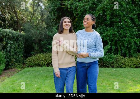 Weibliche Freunde, die Arm in Arm auf dem Rasen laufen, tragen lässige Pullover und Jeans im Park Stockfoto