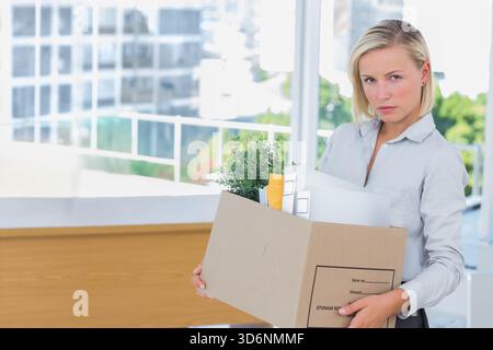 Frau in Bluse und Hose, die Pappkarton mit Pflanze hält, Ordner an der Rezeption, Kopierraum. Umzug, Arbeitsplatz, Urban, Glas, minimalistisch, Stockfoto