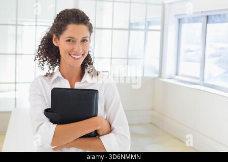 Erwachsene Frau, die in einem hellen Büro mit Gitterfenstern steht und ein weißes Hemd trägt. Professionell, Arbeitsbereich, Ordner, minimalistisch, Modus Stockfoto