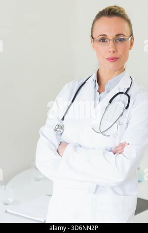 Arztin mittleren Alters mit gekreuzten Armen in der Klinik mit Labormantel und Stethoskop. Minimalistisch, professionell, medizinisch, Büro, ruhig, c Stockfoto