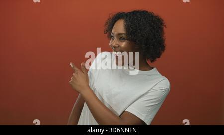 Frau mit lockigen Haaren lächelt und zeigt mit dem Finger im Studio-Porträt mit weißem T-Shirt; Aufregung. Stockfoto
