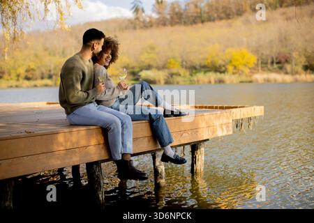Ein fröhliches Paar sitzt nebeneinander auf einem hölzernen Dock, schlürft Drinks und lächelt sich an einem warmen Herbsttag an. Stockfoto