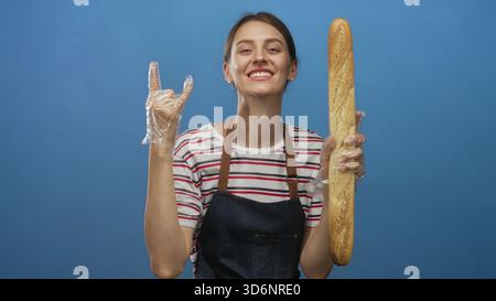 Frau mit Baguette, Schürze und Geste mit Steinhörnern im blauen Studio; verspielte Backenergie. Stockfoto