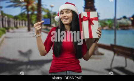 Junge Frau hält Kreditkarten und Geschenkverpackungen, trägt weihnachtsmütze, lächelt und zeigt Karte auf der Straße; Urlaubsfreude. Stockfoto