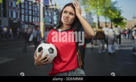Frau, die Fußball mit der Hand auf der Stirn hält, rotes Jersey in einer überfüllten Straße von Gebäuden und Bäumen trägt, eine verwirrte Grimasse ausdrückt; Frustra Stockfoto