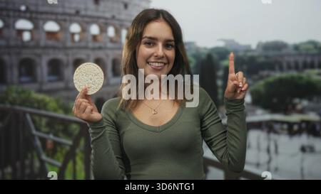 Frau hält einen runden Reiskuchen und zeigt den Finger in das Gebäude in der Nähe des römischen kolosseums, lächelt und präsentiert Snacks; gesunde Snackfreude. Stockfoto