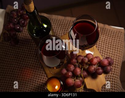 Nahaufnahme von oben zwei Weingläsern mit Rotwein, Flasche auf einem Tisch, Traubenbündel in der Nähe romantischer Kerzen mit brennendem Feuer. Konzept von Chris Stockfoto