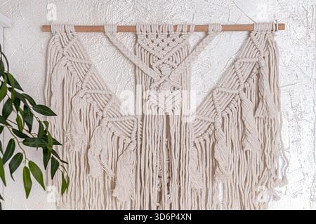 Makramee-Wandbehang im Boho-Stil, handgefertigt aus Baumwollseil, ziert eine strukturierte helle Wand. Trendige moderne Wohnkultur, Bastelarbeit, gemütliche Stimmung. Stockfoto