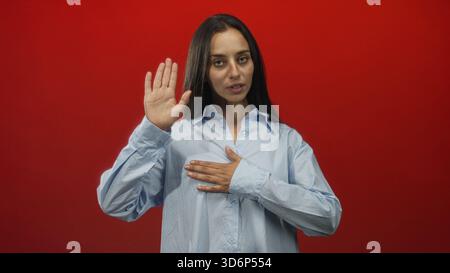 Frau hebt die Hand in die Kamera im roten Studio, während sie die andere Hand als Eid-Geste auf die Brust legt; feierliches Pfand. Stockfoto
