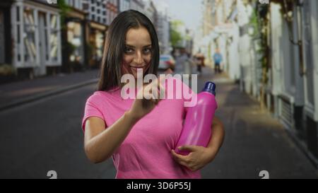 hispanische junge Frau, die eine rosa Flasche in einem Arm hält und mit dem Zeigefinger auf eine Stadtstraße zeigt, während sie lächelt; verspielte Beförderung. Stockfoto