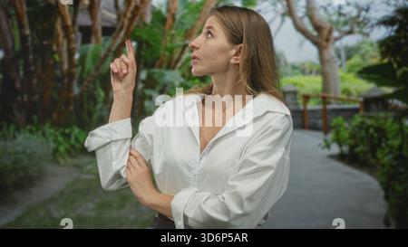 Die Frau zeigt den Finger nach oben auf einem Waldparkweg, trägt ein weißes Hemd mit gerollten Ärmeln und gefalteten Armen und blickt nach oben; Neugier entdeckt. Stockfoto