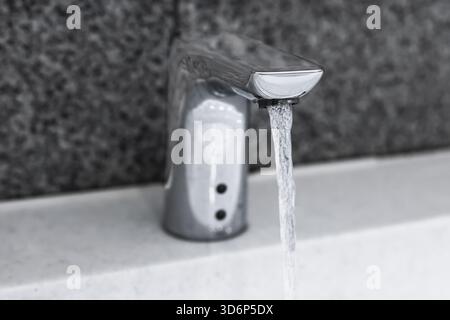 Ein Wasserstrahl, der von einem modernen, automatischen Sensorhahn aus Chrom über eine weiße Ablagefläche fließt. Hochwertige Fotos Stockfoto