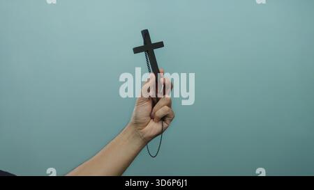 Männliche Hand, die einen schwarzen Kreuz-Anhänger mit Kette vor einer blauen Hintergrundwand hält, symbolisiert Glauben, Hingabe und religiösen Glauben in einer isolierten se Stockfoto