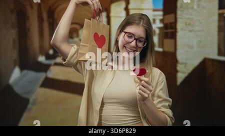Frau in Brille hält Paperbag mit rotem Herz und Herzausschnitt im Gebäudeflur, während sie lächelt; Liebesfreude. Stockfoto