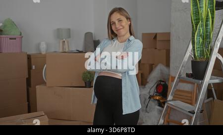 Schwangere Frau lächelt selbstbewusst im Wohnzimmer, umgeben von Umzugskästen, und zeigt einen Neuanfang in ihrem neuen Zuhause mit Pflanzen und Möbeln. Stockfoto