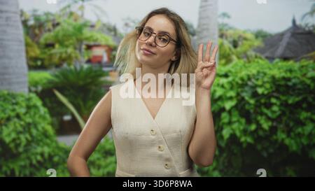 Frau in ärmellosem Kleid, die eine Brille trägt, hält drei Finger hoch und lächelt in einem Waldpark mit Palmen und grünen Hecken; Zuversicht. Stockfoto