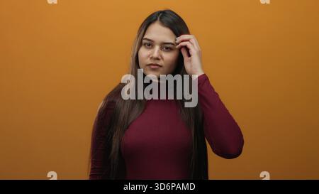 Junge hispanische Frau mit langen Haaren, die mit der Hand vor orangem Hintergrund bürsten, intensiv die Haarspitzen auf das Split-Haarpflegekonzept untersucht. Stockfoto