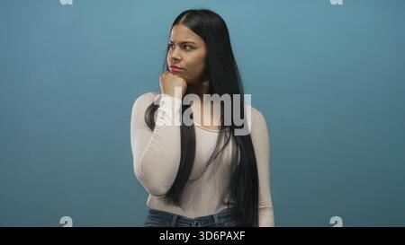 Lateinische Frau posiert mit Hand am Kinn im Studio vor blauer Wand, beugt sich leicht nach vorne und neigt den Kopf; Zweifel. Stockfoto