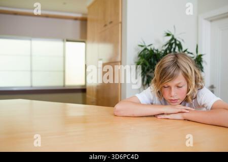 Junge, der zu Hause auf einem großen hellen Holztisch sitzt, weißes T-Shirt trägt, Kopf ruht, Kopierraum. Tageslicht, minimal, gemütlich, Küche, Esszimmer, holzkorn, S Stockfoto
